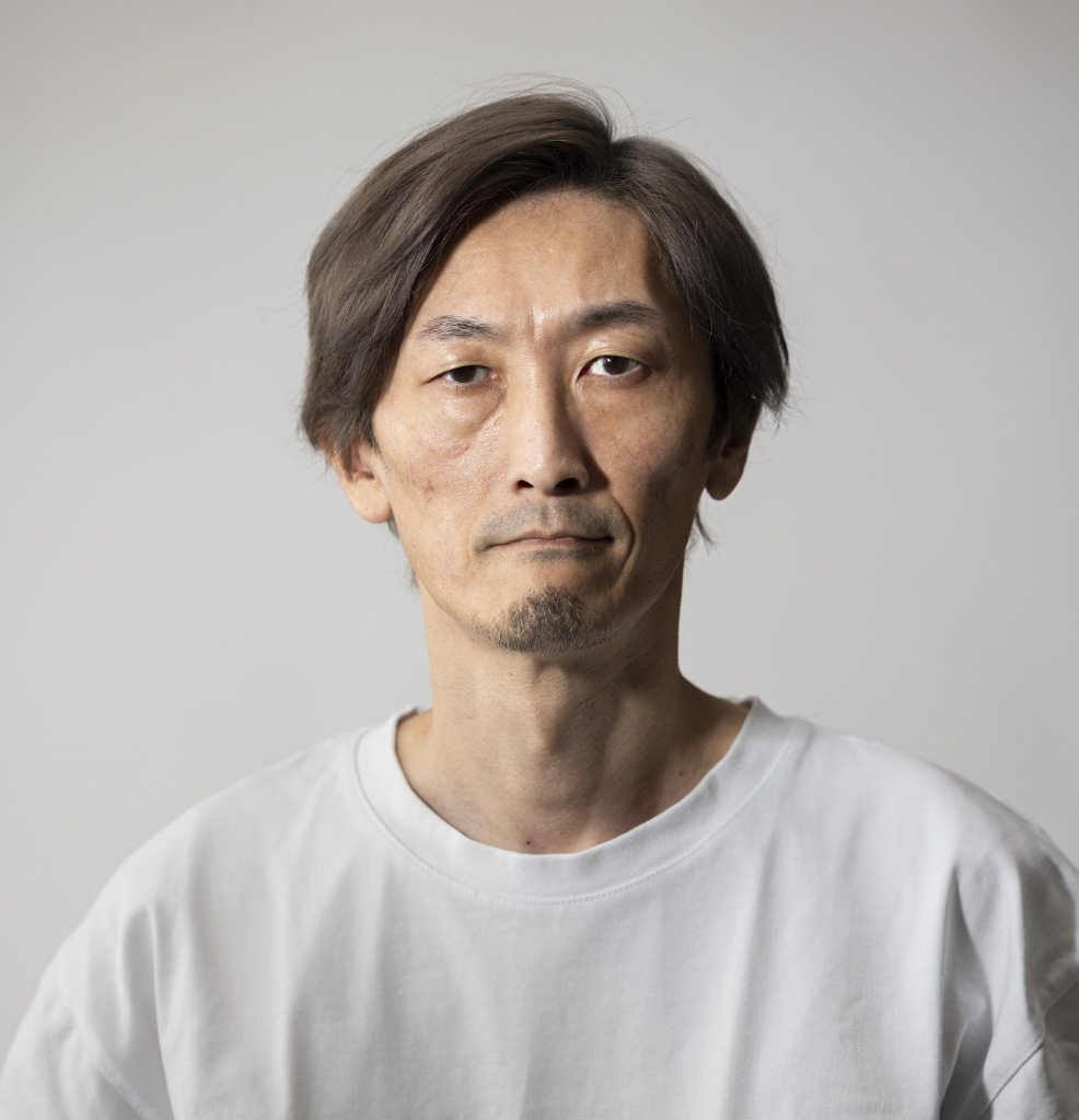 中年男性が正面を向いた写真。表情は真顔で白いTシャツを着ている。